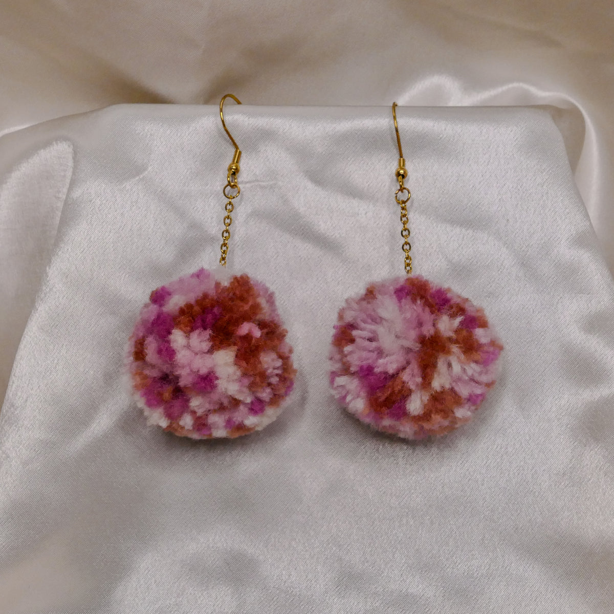 A pair of handmade dangle earrings with fluffy pom-poms in pink, purple, and white, hanging from gold fishhook wires on a white satin fabric background.