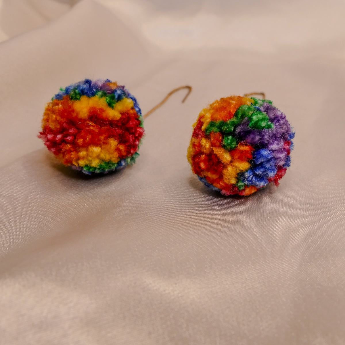 A pair of handmade, colorful pom-pom earrings made from textured yarn, featuring a rainbow of red, orange, yellow, green, blue, and purple colors, resting on a soft beige fabric background.